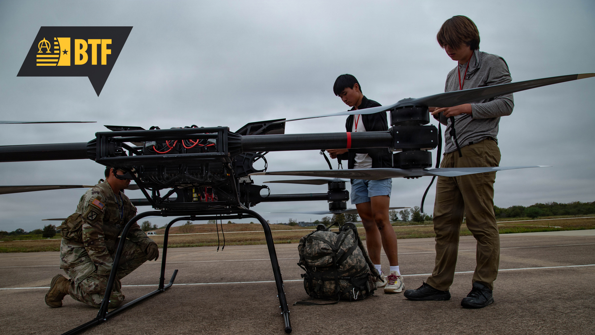 Three individuals work around a large multi‑rotor drone on a paved airfield under an overcast sky. One individual in camouflage uniform kneels near the drone’s landing gear, appearing to inspect equipment. Two other individuals stand on the opposite side, handling cables or components. A large backpack rests on the ground beside the drone. The drone has multiple long rotor blades and exposed wiring at its center. A black and yellow logo with the letters ‘BTF’ appears in the upper left corner. The background shows open fields and low vegetation.