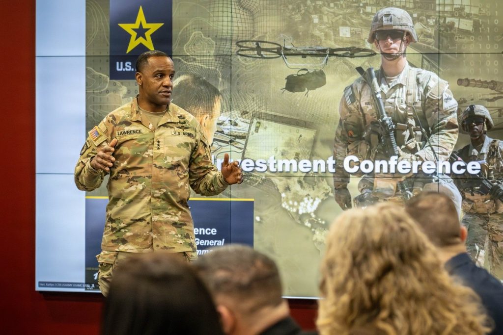 A person in a U.S. Army camouflage uniform stands and speaks in front of a large presentation screen. The screen displays military‑themed graphics, including a soldier in gear, a drone, and the words “Investment Conference.” Several people seated in the foreground face the presenter. The setting appears to be a conference or briefing room with professional military visuals on the display wall.